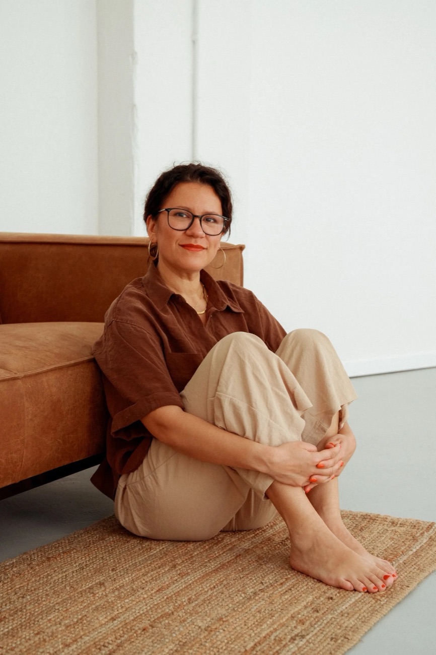 Sophie sitzt entspannt auf dem Boden vor einem Sofa, in brauner Bluse und beiger Hose